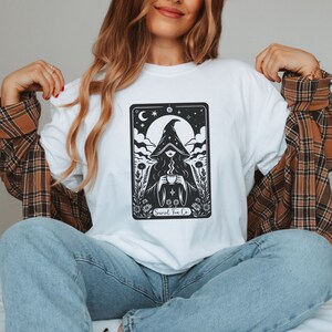 Tarot card design graphic with a mysterious woman wearing a hooded cloak and drinking tea, surrounded by wildflowers, stars and large moon in the sky. Text Suriel Tea Co at the bottom. ACOTAR inspired PNG SVG DXT cut file. Perfect for print on demand