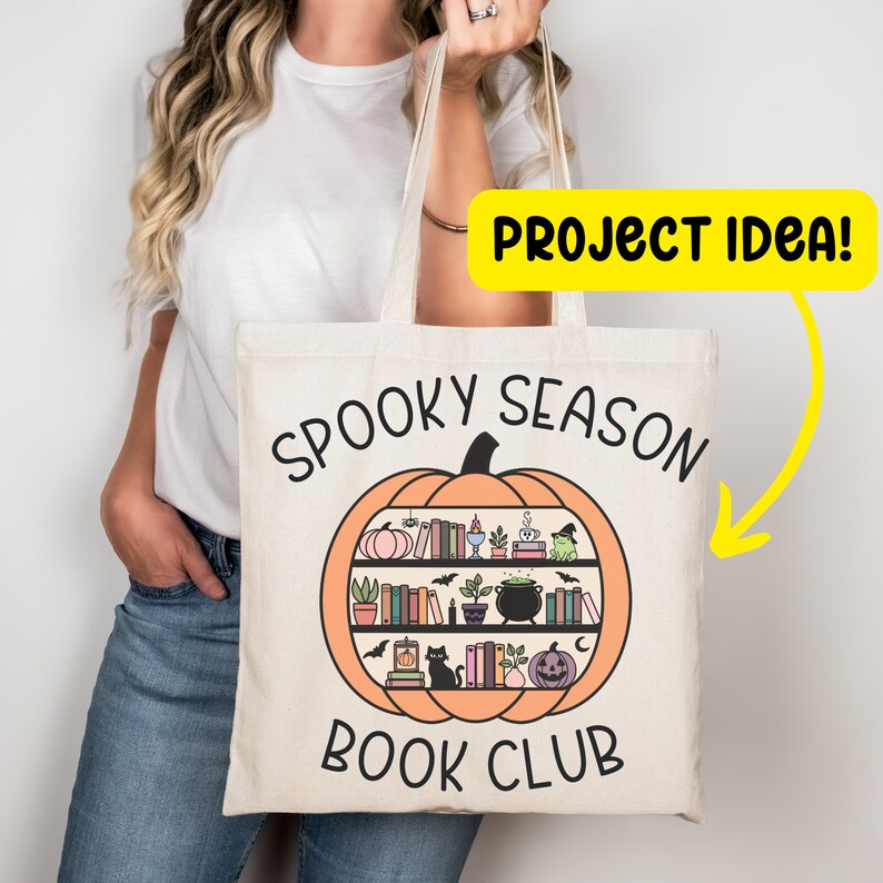 May include: A white tote bag with the text "Spooky Season Book Club" printed on it. The text is surrounded by a pumpkin with a bookshelf inside. The bookshelf is filled with books, plants, and other Halloween-themed items.