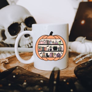 May include: White ceramic mug with a black outline of a pumpkin. The pumpkin is filled with books, a cauldron, a cat, and other Halloween-themed items.