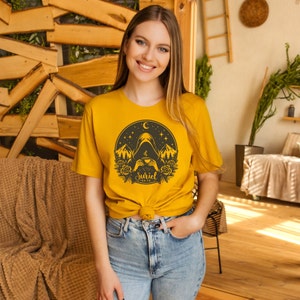 K&ouml;nnte beinhalten: Eine Frau tr&auml;gt ein senfgelbes T-Shirt mit einem schwarz-wei&szlig;en Grafikdesign einer Kapuzenfigur, die eine Tasse Tee h&auml;lt. Das Grafikdesign beinhaltet Berge, Sterne, einen Halbmond und Rosen. Der Text "Suriel Tea Co." ist auf dem Grafikdesign gedruckt.
