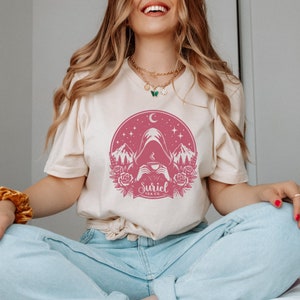 K&ouml;nnte beinhalten: Ein wei&szlig;es T-Shirt mit einem rosafarbenen Grafikdesign einer Kapuzenfigur, die eine Tasse Tee h&auml;lt. Die Grafik enth&auml;lt auch Berge, Sterne, einen Halbmond und den Text "Suriel Tea Co."