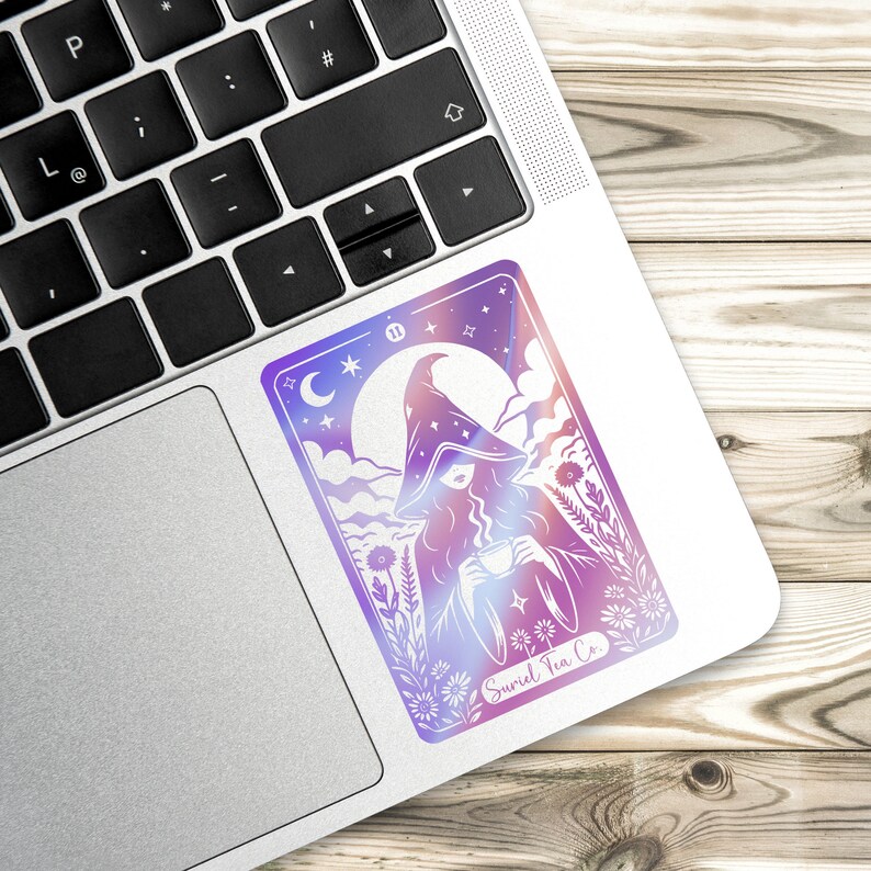 Tarot card design graphic with a mysterious woman wearing a hooded cloak and drinking tea, surrounded by wildflowers, stars and large moon in the sky. Text Suriel Tea Co at the bottom. ACOTAR inspired PNG SVG DXT cut file. Perfect for print on demand