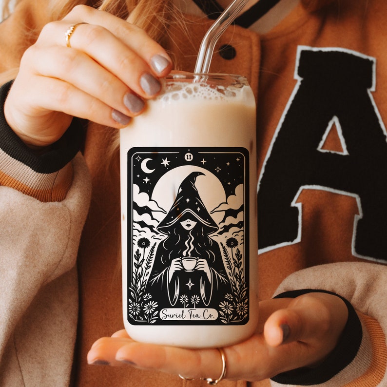 Tarot card design graphic with a mysterious woman wearing a hooded cloak and drinking tea, surrounded by wildflowers, stars and large moon in the sky. Text Suriel Tea Co at the bottom. ACOTAR inspired PNG SVG DXT cut file. Perfect for print on demand
