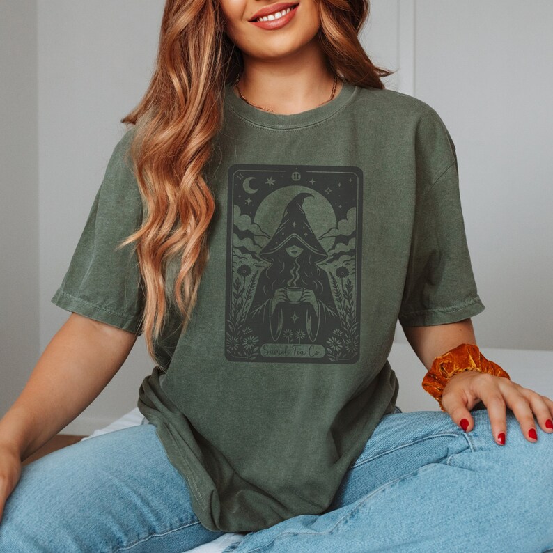 Tarot card design graphic with a mysterious woman wearing a hooded cloak and drinking tea, surrounded by wildflowers, stars and large moon in the sky. Text Suriel Tea Co at the bottom. ACOTAR inspired PNG SVG DXT cut file. Perfect for print on demand