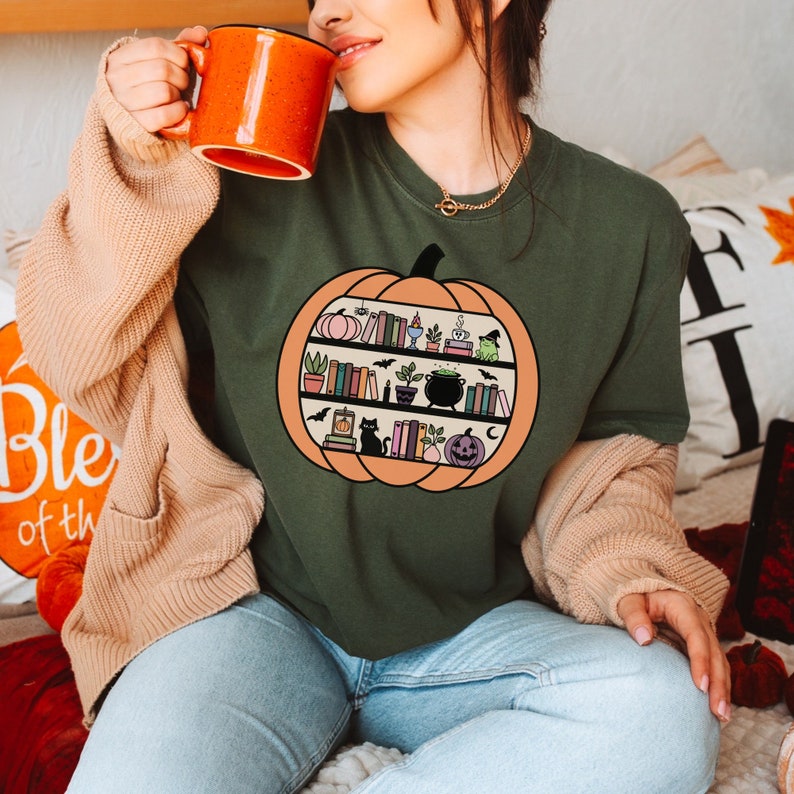 May include: A green t-shirt with a graphic of a pumpkin filled with books, a cauldron, a frog, and other Halloween-themed items. The text on the shirt says "Bookish & Spooky".