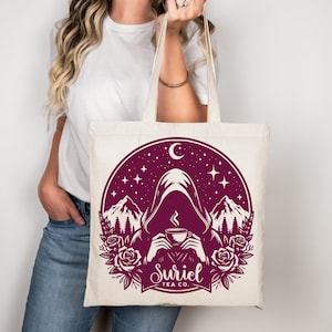 K&ouml;nnte beinhalten: Eine wei&szlig;e Canvas-Tragetasche mit einem burgunderfarbenen Design einer Kapuzenfigur, die eine dampfende Tasse Tee h&auml;lt. Das Design ist vor dem Hintergrund von Bergen, Sternen und einem Halbmond platziert. Der Text "Suriel Tea Co." ist unter der Figur gedruckt.