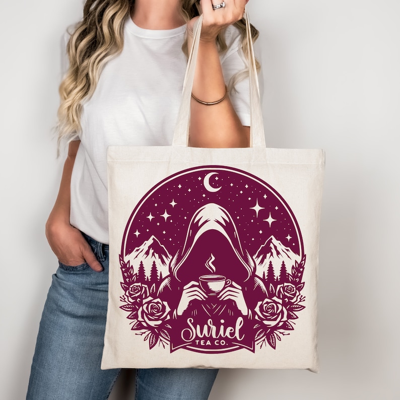 K&ouml;nnte beinhalten: Eine wei&szlig;e Canvas-Tragetasche mit einem burgunderfarbenen Design einer Kapuzenfigur, die eine dampfende Tasse Tee h&auml;lt. Das Design ist vor dem Hintergrund von Bergen, Sternen und einem Halbmond platziert. Der Text "Suriel Tea Co." ist unter der Figur gedruckt.