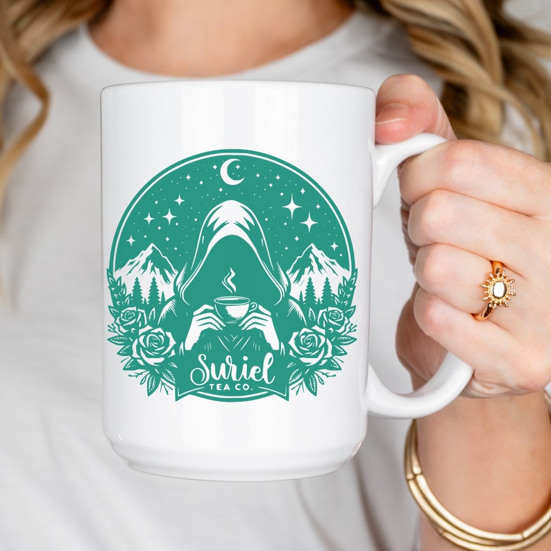 K&ouml;nnte beinhalten: Eine wei&szlig;e Keramiktasse mit einem blaugr&uuml;nen Design einer Kapuzenfigur, die eine Tasse Tee h&auml;lt. Das Design beinhaltet Berge, Sterne und den Text "Suriel Tea Co."