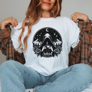 K&ouml;nnte beinhalten: Wei&szlig;es T-Shirt mit einem schwarz-wei&szlig;en Grafikdesign einer Kapuzenfigur, die eine Tasse Tee h&auml;lt. Die Grafik enth&auml;lt einen Halbmond, Sterne, Berge und Rosen. Der Text "Suriel Tea Co." befindet sich unter der Figur.