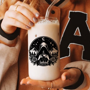 K&ouml;nnte beinhalten: Ein klares Glas mit einer schwarz-wei&szlig;en Grafik einer Kapuzenfigur, die eine Tasse in einer Waldlandschaft h&auml;lt. Die Grafik enth&auml;lt den Text "Suriel Tea Co.". Das Glas ist mit einem hellbraunen Getr&auml;nk gef&uuml;llt.