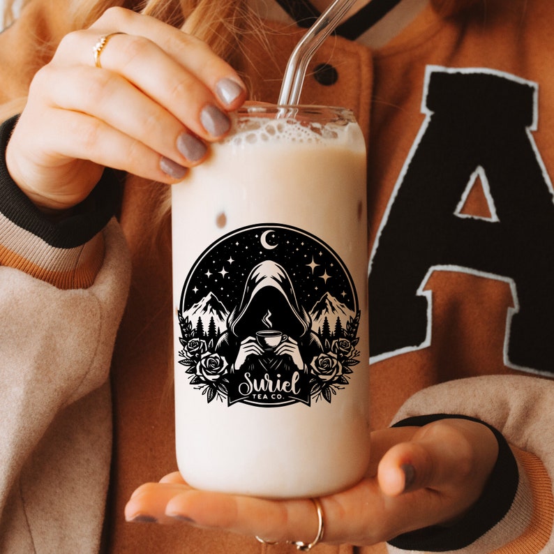 K&ouml;nnte beinhalten: Ein klares Glas mit einer schwarz-wei&szlig;en Grafik einer Kapuzenfigur, die eine Tasse in einer Waldlandschaft h&auml;lt. Die Grafik enth&auml;lt den Text "Suriel Tea Co.". Das Glas ist mit einem hellbraunen Getr&auml;nk gef&uuml;llt.