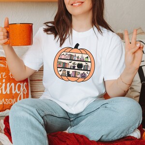 May include: White t-shirt with a graphic of a pumpkin filled with books, a cauldron, a black cat, and other Halloween-themed items.