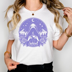 K&ouml;nnte beinhalten: Wei&szlig;es T-Shirt mit einem lilafarbenen Design einer Kapuzenfigur, die eine Tasse Tee vor einer Berglandschaft mit Sternen und einem Halbmond h&auml;lt. Das Design enth&auml;lt auch den Text "Suriel Tea Co."