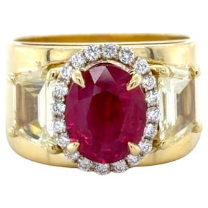 Puede incluir: Un anillo de oro con una gran piedra central de rubí ovalada, rodeada de pequeños diamantes. El anillo tiene dos piedras laterales rectangulares de color claro y una banda ancha de oro pulido. El rubí es de color rojo intenso y los diamantes brillan.