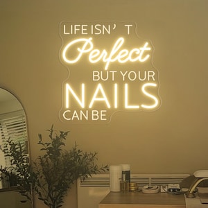 Könnte beinhalten: Ein weißes Neon-Schild mit dem Text "Life isn't perfect but your nails can be" hängt an einer beigefarbenen Wand in einem Nagelstudio. Das Schild leuchtet und die Buchstaben sind in einer kursivschrift.