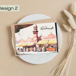 K&ouml;nnte beinhalten: Aquarell-Gru&szlig;karte mit einer Moschee mit Minarett und Marktst&auml;nden. Die Karte tr&auml;gt den Text "Design 2" und arabische Schriftzeichen. Die Karte liegt auf einem wei&szlig;en Teller mit getrockneten Blumen.
