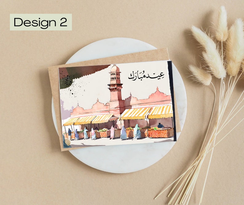 K&ouml;nnte beinhalten: Aquarell-Gru&szlig;karte mit einer Moschee mit Minarett und Marktst&auml;nden. Die Karte tr&auml;gt den Text "Design 2" und arabische Schriftzeichen. Die Karte liegt auf einem wei&szlig;en Teller mit getrockneten Blumen.