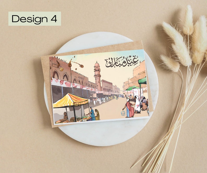 K&ouml;nnte beinhalten: Aquarell-Gru&szlig;karte mit einer Stra&szlig;enszene, Moschee und Marktst&auml;nden. Der Text "Design 4" und arabische Schrift sind zu sehen. Die Karte liegt auf einem wei&szlig;en Teller, begleitet von getrockneten Blumen.