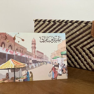 K&ouml;nnte beinhalten: Eine Gru&szlig;karte im Aquarellstil mit einer gesch&auml;ftigen Stra&szlig;enszene mit Moschee und Marktst&auml;nden. Die Karte hat arabische Kalligraphie und liegt neben einem geflochtenen Korb mit Lederverschluss.