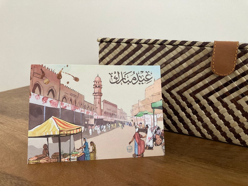 K&ouml;nnte beinhalten: Eine Gru&szlig;karte im Aquarellstil mit einer gesch&auml;ftigen Stra&szlig;enszene mit Moschee und Marktst&auml;nden. Die Karte hat arabische Kalligraphie und liegt neben einem geflochtenen Korb mit Lederverschluss.