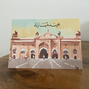 K&ouml;nnte beinhalten: Gru&szlig;karte mit einer Aquarellillustration eines gro&szlig;en Geb&auml;udes mit gew&ouml;lbten T&uuml;ren und einem zentralen Eingang. Die Karte hat arabischen Text oben und ist vor einem hellbraunen und gelben Hintergrund.