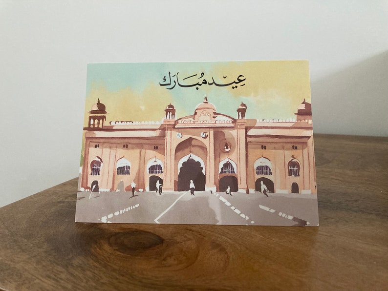 K&ouml;nnte beinhalten: Gru&szlig;karte mit einer Aquarellillustration eines gro&szlig;en Geb&auml;udes mit gew&ouml;lbten T&uuml;ren und einem zentralen Eingang. Die Karte hat arabischen Text oben und ist vor einem hellbraunen und gelben Hintergrund.