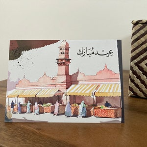 K&ouml;nnte beinhalten: Eine Aquarell-Gru&szlig;karte mit einer nah&ouml;stlichen Marktszene mit einer Moschee und Figuren in traditioneller Kleidung. Die Karte hat arabischen Text oben. Die Farbpalette umfasst Rosa-, Braun- und Gelbt&ouml;ne.