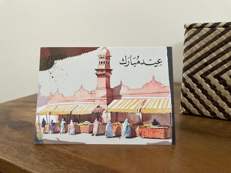 K&ouml;nnte beinhalten: Eine Aquarell-Gru&szlig;karte mit einer nah&ouml;stlichen Marktszene mit einer Moschee und Figuren in traditioneller Kleidung. Die Karte hat arabischen Text oben. Die Farbpalette umfasst Rosa-, Braun- und Gelbt&ouml;ne.