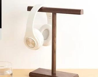 May include: A wooden headphone stand with a dark brown finish. The stand has a T-shaped top with a single arm extending down to a rectangular base. A white pair of headphones is hanging on the stand.