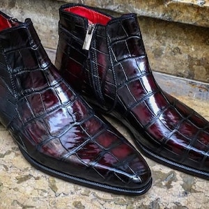 May include: A pair of burgundy leather ankle boots with a crocodile skin pattern. The boots have a zipper closure on one side and a buckle on the other. The interior lining is red.