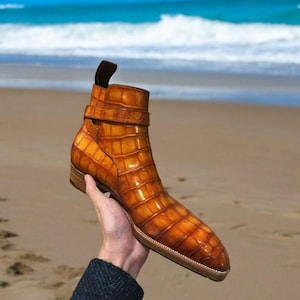 May include: A pair of brown leather ankle boots with a crocodile pattern. The boots have a buckle strap and a pointed toe.