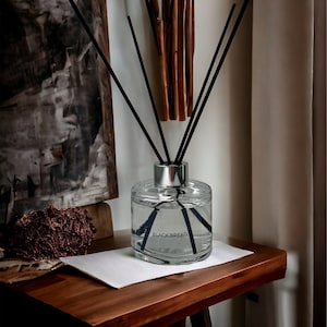 Puede incluir: Una botella difusora de vidrio transparente con cañas negras y una tapa plateada. La botella está llena de un líquido transparente y tiene una etiqueta que dice "Black Bread".