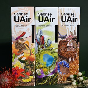 Puede incluir: Tres cajas de ambientador con el nombre de la marca "Sabrise UAir" impreso en la parte delantera. Las cajas presentan diferentes diseños: una libélula, un paisaje verde y una hogaza de pan. El texto en las cajas dice "кохайтеся", "зелені свята" y "чорний хліб".
