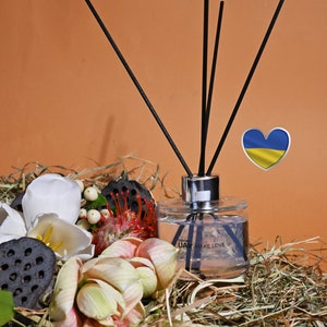 May include: A clear glass bottle with black diffuser sticks and a label that reads "UAIR MAKE LOVE". The bottle is sitting on a bed of dried grass with a white flower, a red flower, and a dried lotus pod. A heart-shaped sticker with the Ukrainian flag is in the background.