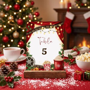 Könnte beinhalten: Ein rechteckiges Tischnummernschild mit der Aufschrift Table 5 im Kinodesign mit Filmstreifen, roten Vorhängen, einem Popcornbecher und einem Becher mit heißer Schokolade. Das Motiv enthält weihnachtliches Grün und Lichterketten auf einem Holzsockel.