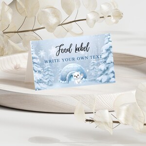 May include: A rectangular folded paper food label featuring a winter scene with a white polar bear standing in front of an igloo, surrounded by snow-covered trees and snowflakes. The text reads Food label and WRITE YOUR OWN TEXT.