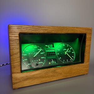 Könnte beinhalten: Eine rechteckige Holzuhr mit integriertem Auto-Instrumentenbrett und grün beleuchtetem Display. Die Anzeige umfasst einen Tachometer in km/h, eine Uhr und einen digitalen Kilometerzähler mit dem Wert 27195, eingefasst in Eichenholz.