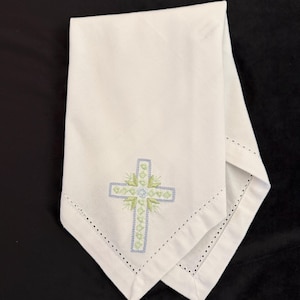May include: White linen napkin with a decorative embroidered cross in light blue and green. The cross is centered on the napkin and surrounded by a floral design. The edges of the napkin are finished with a delicate, dotted hem.