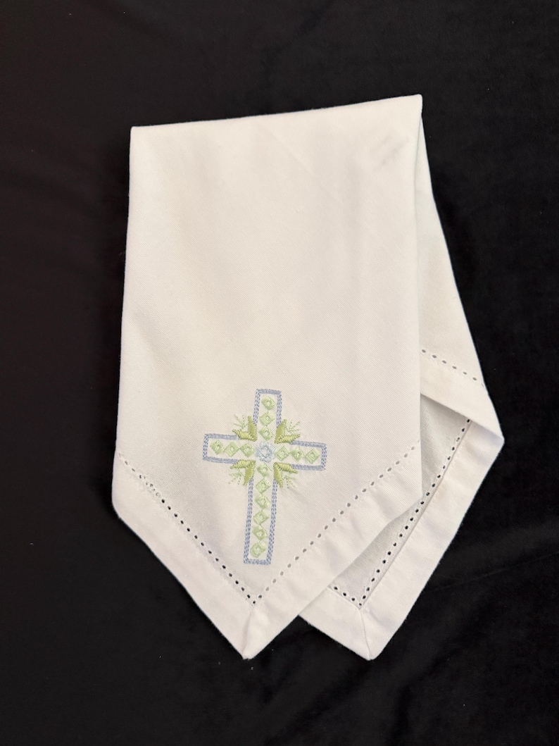 May include: White linen napkin with a decorative embroidered cross in light blue and green. The cross is centered on the napkin and surrounded by a floral design. The edges of the napkin are finished with a delicate, dotted hem.