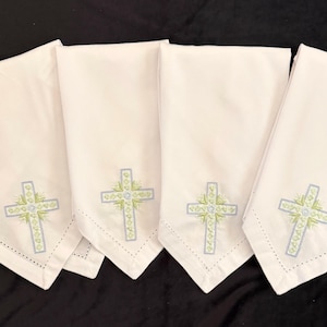 May include: Four white cloth napkins with embroidered crosses. Each napkin features a cross design in light blue and green, with a delicate floral pattern. The napkins are folded and arranged on a black surface.