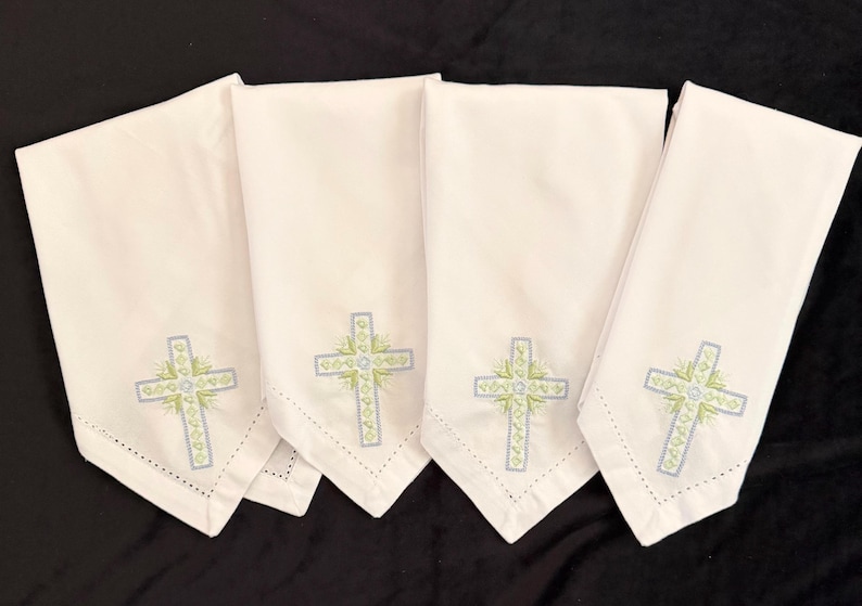 May include: Four white cloth napkins with embroidered crosses. Each napkin features a cross design in light blue and green, with a delicate floral pattern. The napkins are folded and arranged on a black surface.