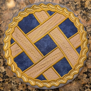 May include: A round, embroidered coaster resembling a pie. The coaster features a blue center with tan lattice strips and a gold scalloped edge. The outer edge is gray.
