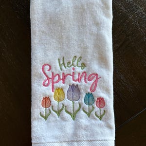 Puede incluir: Toalla de mano blanca bordada con las palabras "Hello Spring" en verde y rosa. Debajo del texto hay tulipanes bordados en tonos rojos, amarillos, morados y azules. La toalla es de material suave y absorbente.