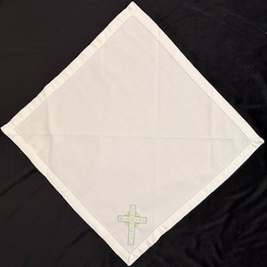 May include: White square linen napkin with a decorative cross embroidered in the lower center. The cross is light blue and green with a detailed design. The edges of the napkin are finished with a dotted stitch.