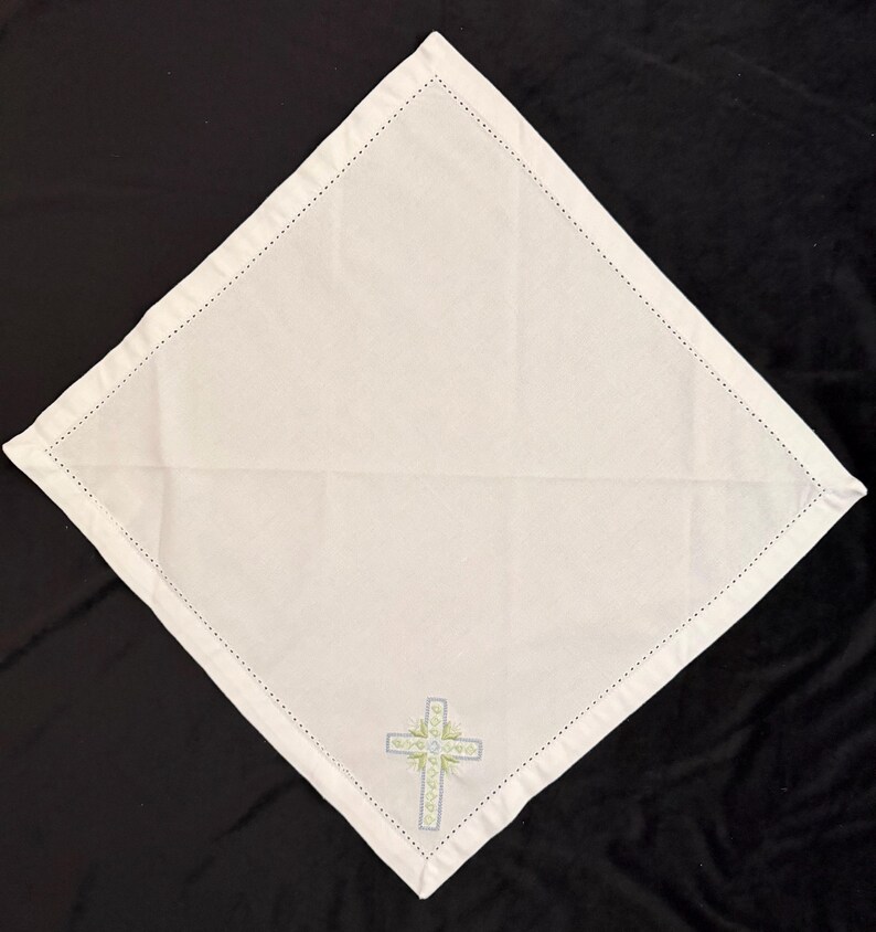 May include: White square linen napkin with a decorative cross embroidered in the lower center. The cross is light blue and green with a detailed design. The edges of the napkin are finished with a dotted stitch.