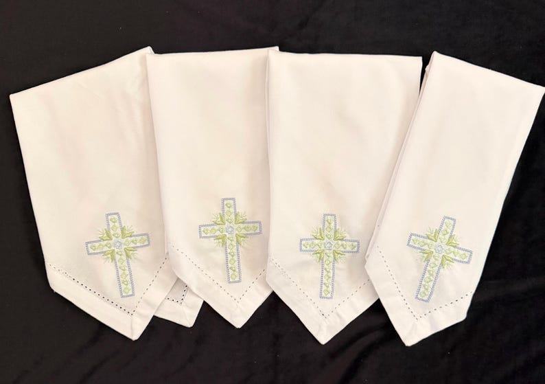 May include: Four white cloth napkins with embroidered crosses. Each napkin features a cross design in light blue and green, with a delicate floral pattern. The napkins are folded and arranged on a black surface.
