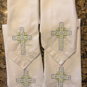 May include: Four white cloth napkins with embroidered crosses. Each napkin features a cross design in light blue and green thread, with a decorative floral element at the center. The napkins have a pointed bottom edge and a stitched border.