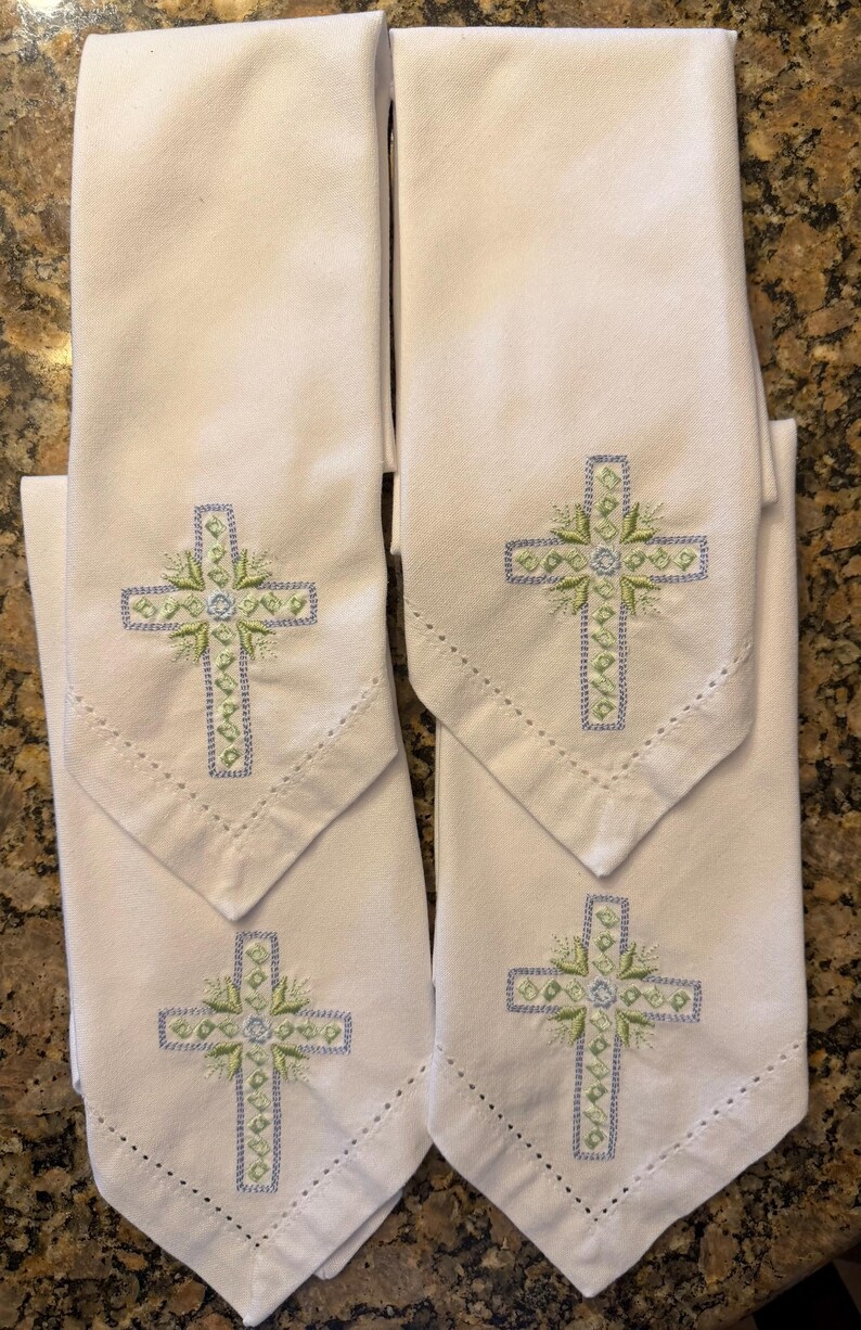 May include: Four white cloth napkins with embroidered crosses. Each napkin features a cross design in light blue and green thread, with a decorative floral element at the center. The napkins have a pointed bottom edge and a stitched border.