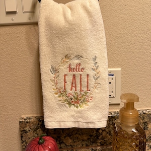 Puede incluir: Toalla de mano color crema con una corona floral bordada y las palabras "hello FALL" en rojo. La toalla está colgada de un anillo de metal negro. Una calabaza roja decorativa y un dispensador de jabón de vidrio están en primer plano.
