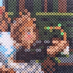 May include: Close-up of a pixel art image made with colorful beads. The image depicts a person's face with brown hair and blue eyes. The background is a mix of green, brown, and black beads.